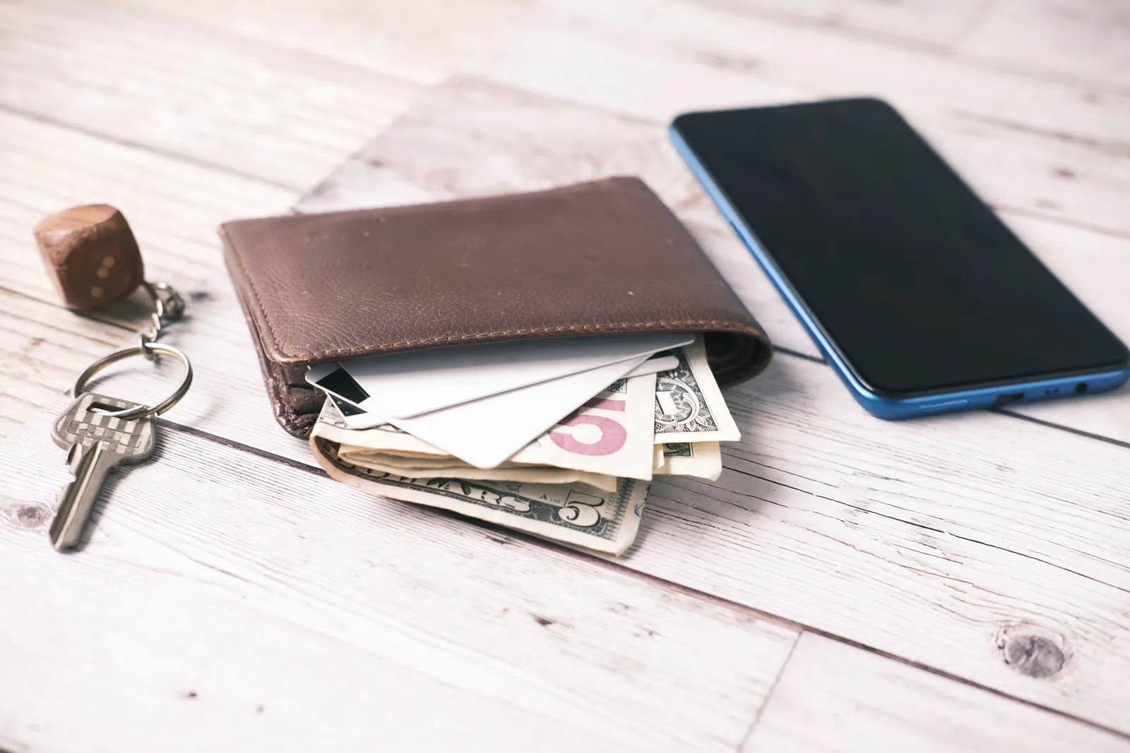 A wallet on a wooden table with cash and cards sticking out, next to a smartphone and a set of keys
